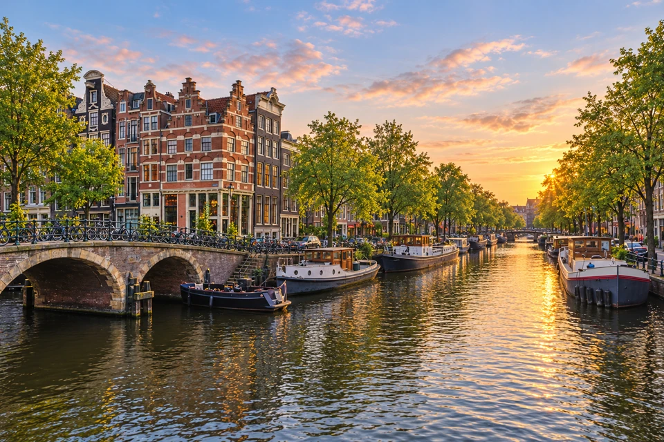 Amsterdam canal with moored barges, a low masonry arch bridge, gabled houses and leafy quays in evening light.