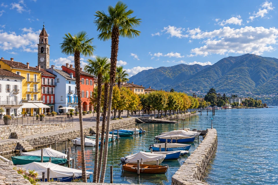 Ascona's colorful lakefront houses and stone bell tower beside palms, covered harbor boats and Lake Maggiore.