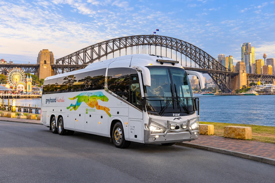 A white three-axle Greyhound Australia coach with a colorful running-dog graphic beside Sydney Harbour.