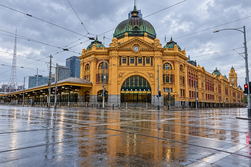 Flinders Street Station's ochre facade and green dome above wet tram tracks in Melbourne.