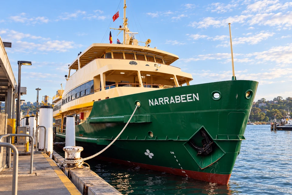 The Sydney ferry Narrabeen moored at a wharf, with a broad green bow, cream wheelhouse, white name and waratah emblem.