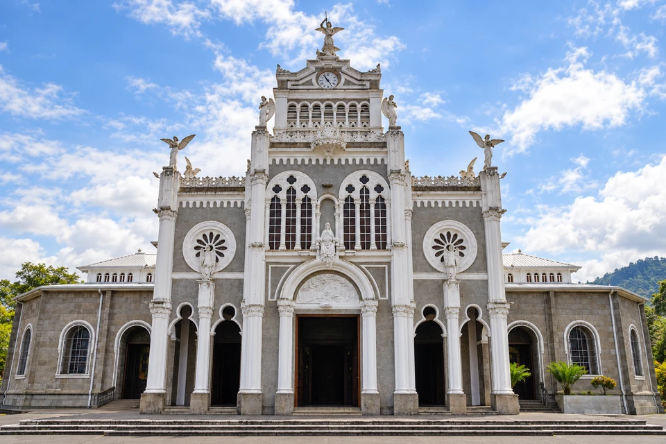 The gray and white facade of Our Lady of the Angels Basilica in Cartago, with rose windows and angel statues.