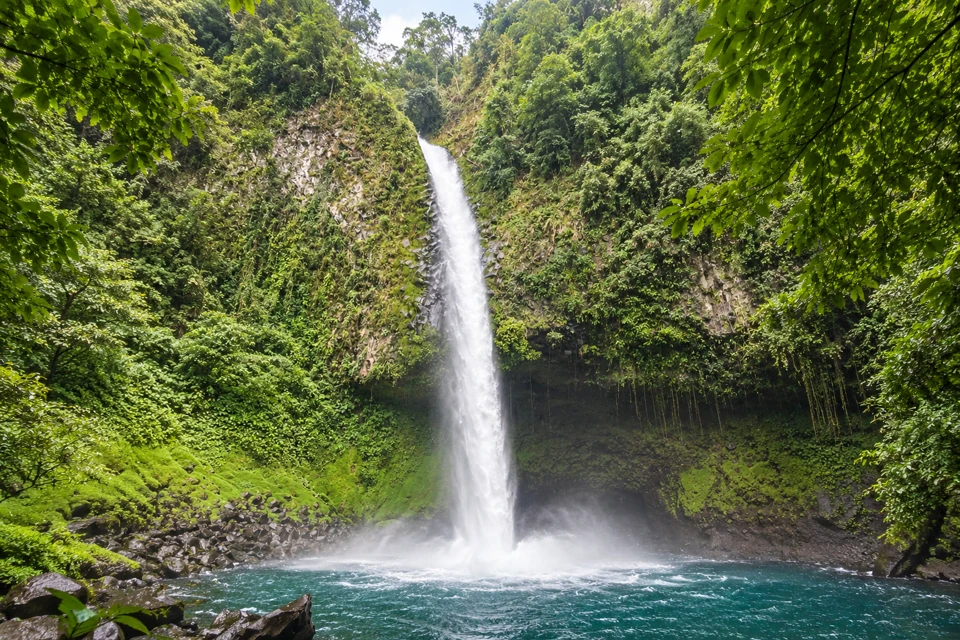 La Fortuna's narrow waterfall dropping over a fern-covered volcanic cliff into a blue-green pool.