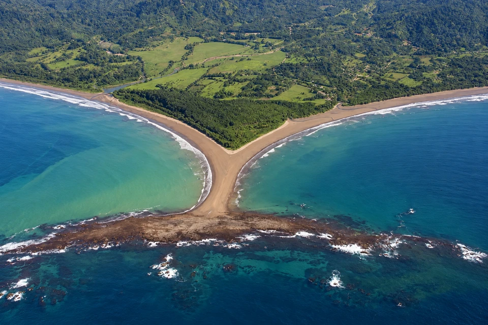 Exposed sand neck and broad rocky reef of the Uvita tombolo, with two bays and forested hills behind.