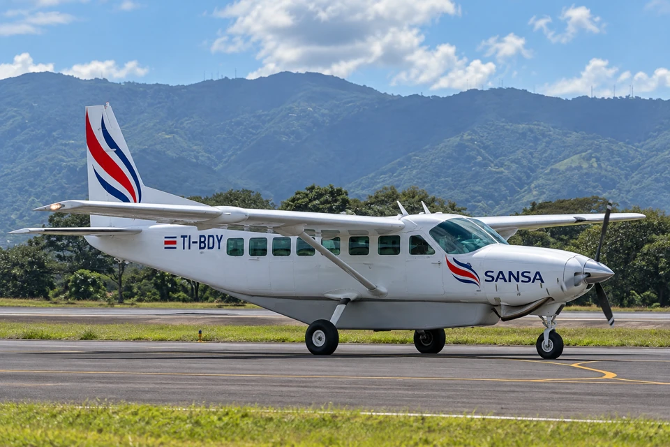 A white SANSA Cessna Grand Caravan with red and blue tail markings, high wings and fixed landing gear at Juan Santamaria Airport.