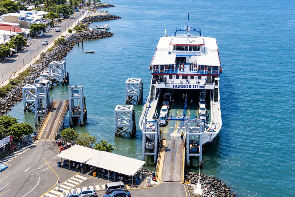 Naviera Tambor's white and blue Tambor III at the Puntarenas vehicle ramp, with cars on its open deck beside Gulf of Nicoya water.