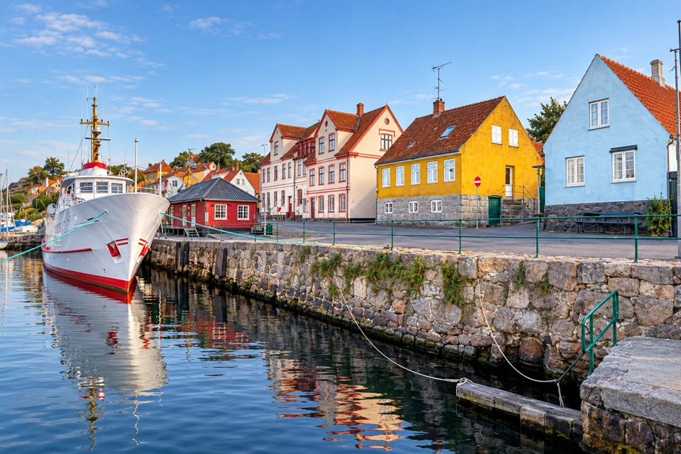 Gudhjem harbour on Bornholm with a granite quay, green railings, colourful red-roofed houses and a moored passenger boat.