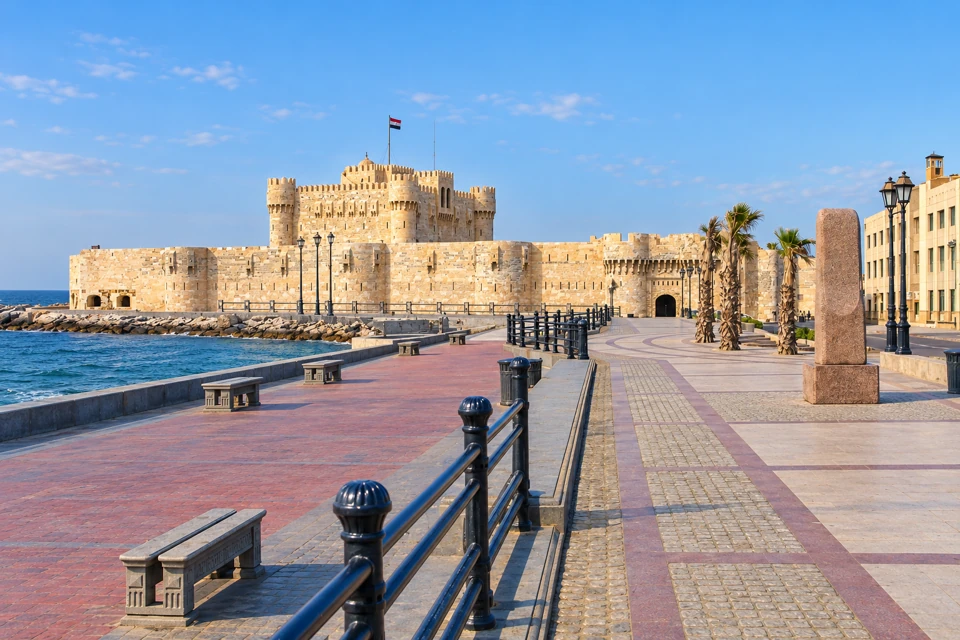 The paved Alexandria waterfront and Mediterranean beside the limestone walls and towers of Qaitbay Citadel.