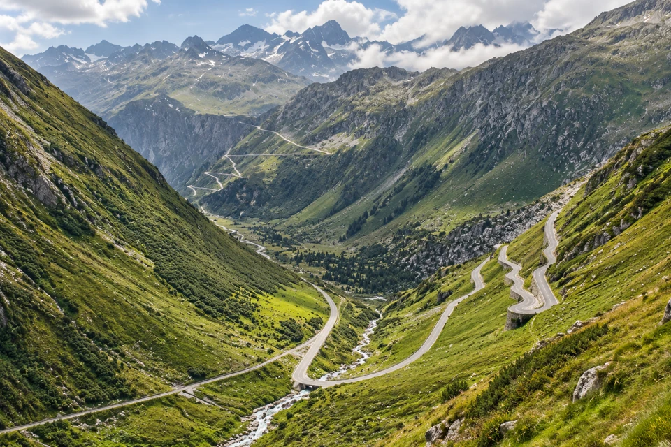 Furka Pass road hairpins on steep green and rocky Alpine valley slopes, with a stream below and craggy ridges beyond.
