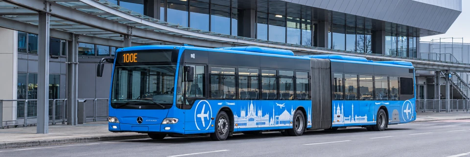 Blue articulated 100E Airport Express bus outside Budapest Airport's glass terminal.