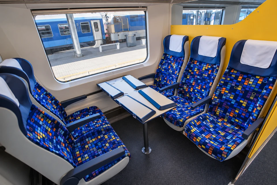 Blue multicolored-square seats with white headrest covers facing across folding table panels in an IC+ coach.