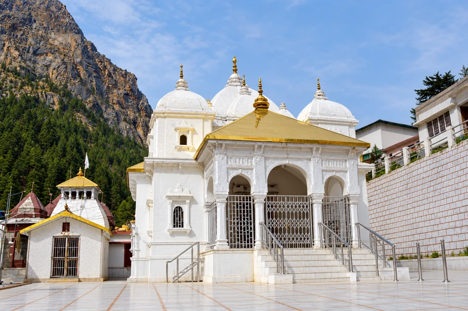 White Gangotri Temple with clustered domed roofs, gold porch roof, metal entry gates and rocky forested hillside