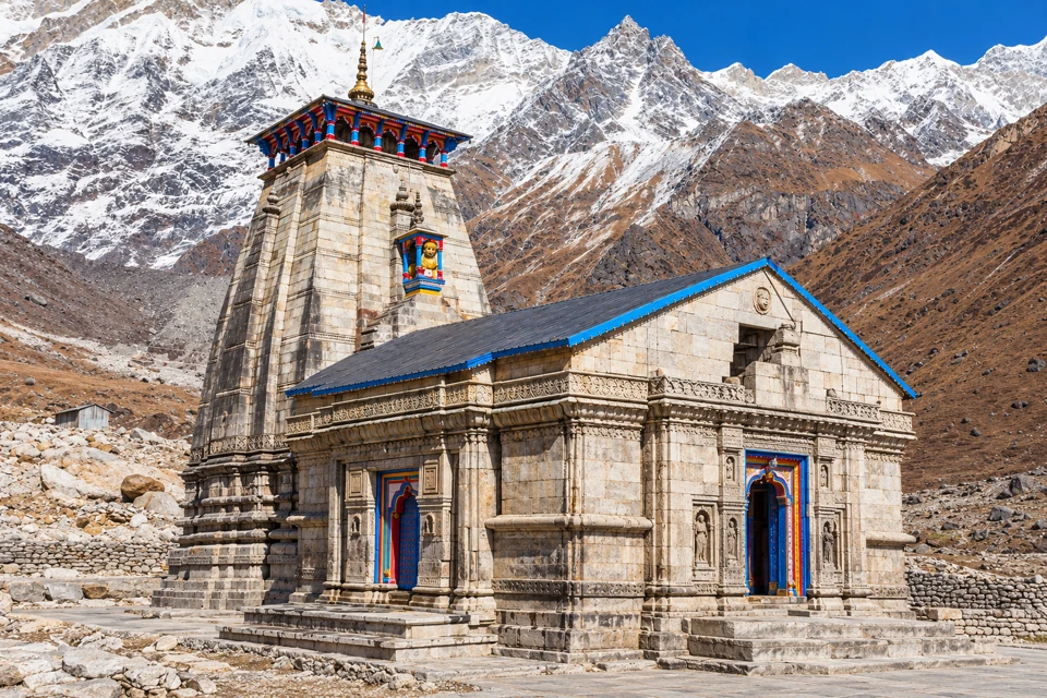 Kedarnath Temple's grey stone tower and hall with blue roof edges, backed by the snowy Himalayan ridge.
