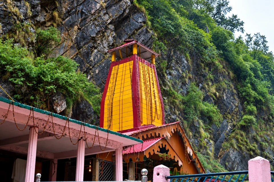 Yamunotri Temple's tapering yellow tower with red bands, open top canopy and pink porch against a steep green rock face