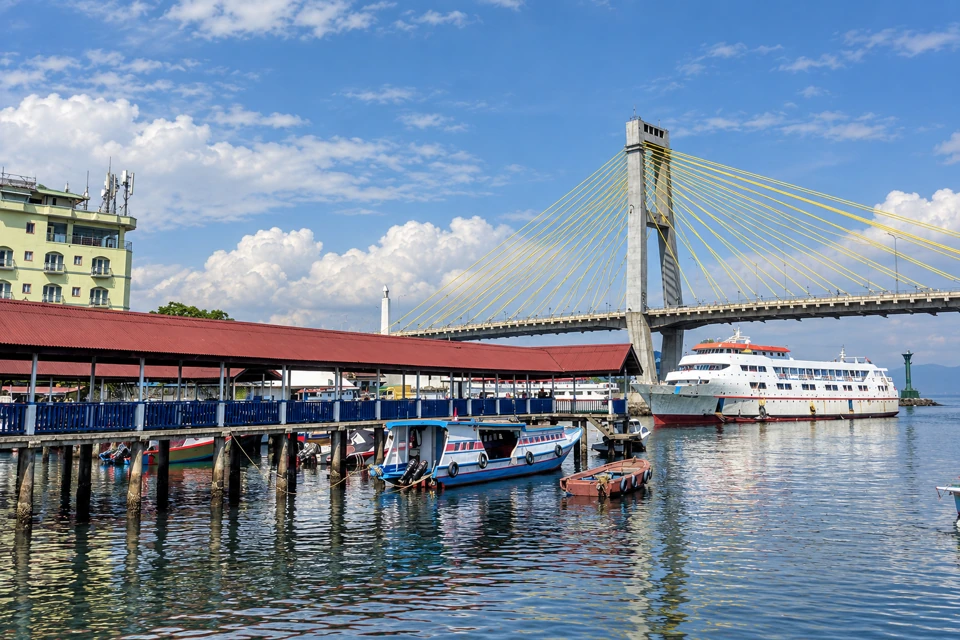 Manado harbor with a red-roofed pier, small passenger boats and Soekarno Bridge's yellow cable fan.