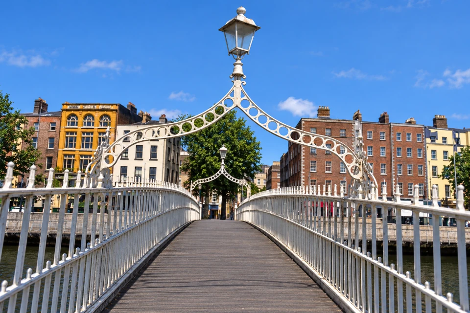 A view along Dublin's Ha'penny Bridge, with pale iron railings, a decorative curved lamp arch and historic buildings beyond.
