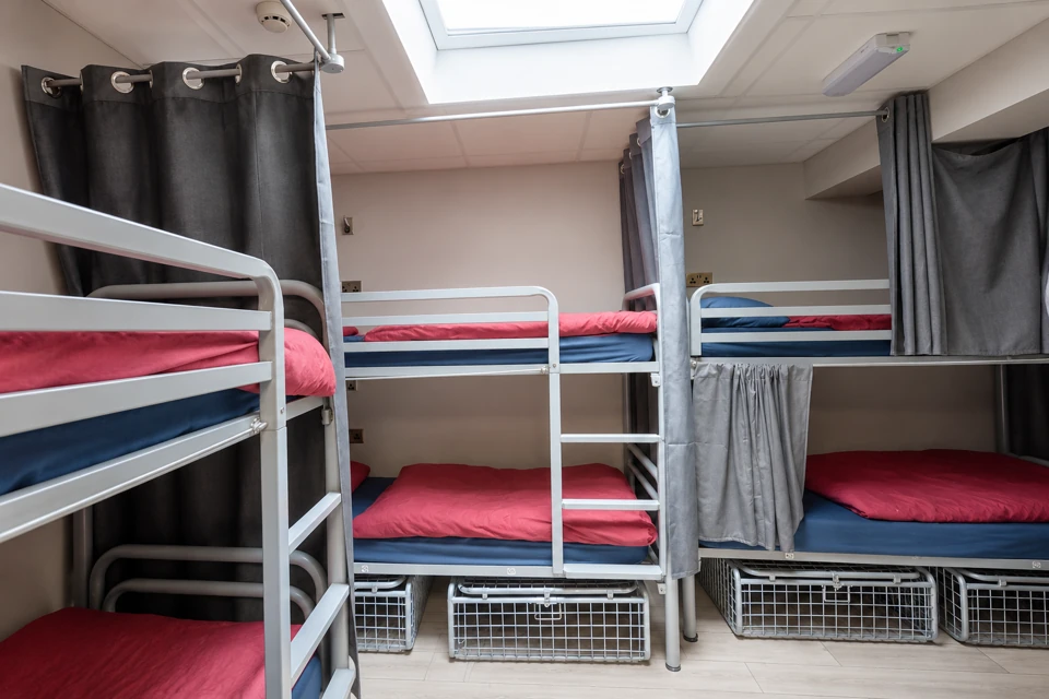 A compact shared Dublin hostel dorm with gray metal bunk beds, red duvets, blue mattresses, privacy curtains and wire storage cages.