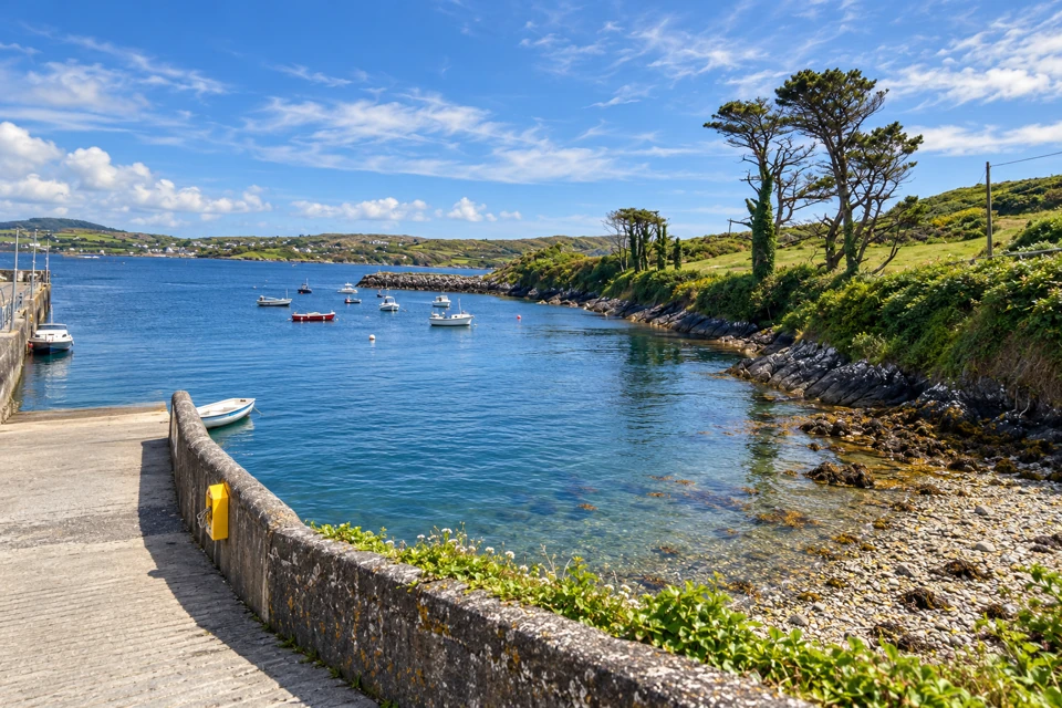 Sherkin Island's sloping stone-edged landing pier, sheltered cove, small moored boats and wind-shaped shoreline trees.