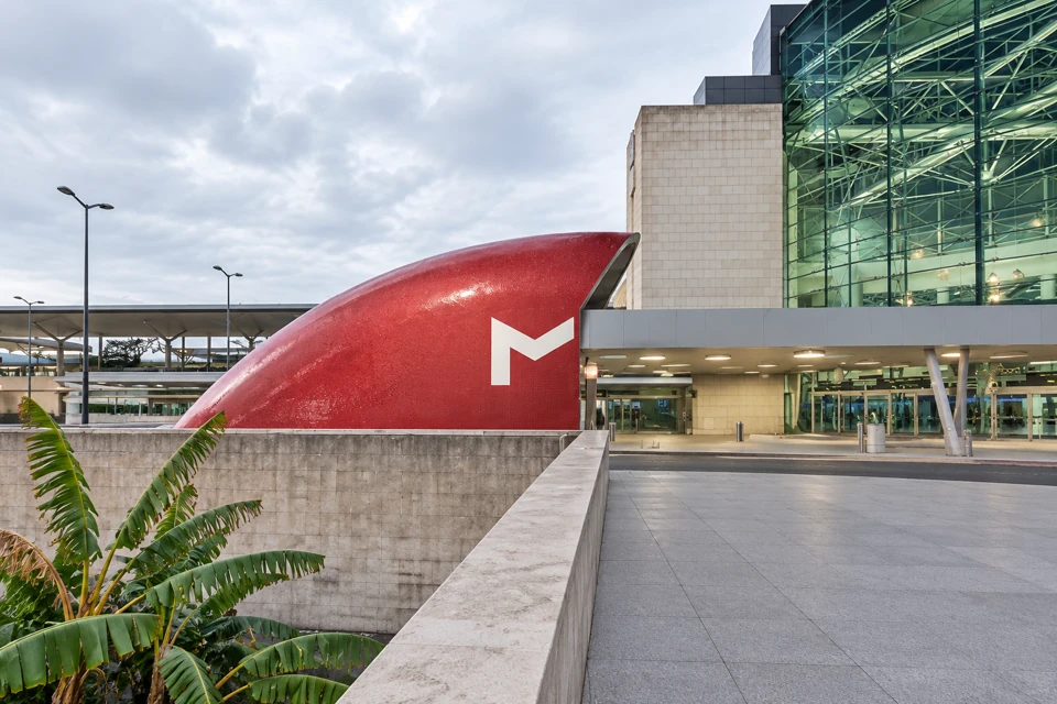 The curved red Metro entrance beside Lisbon Airport's glass terminal frontage.