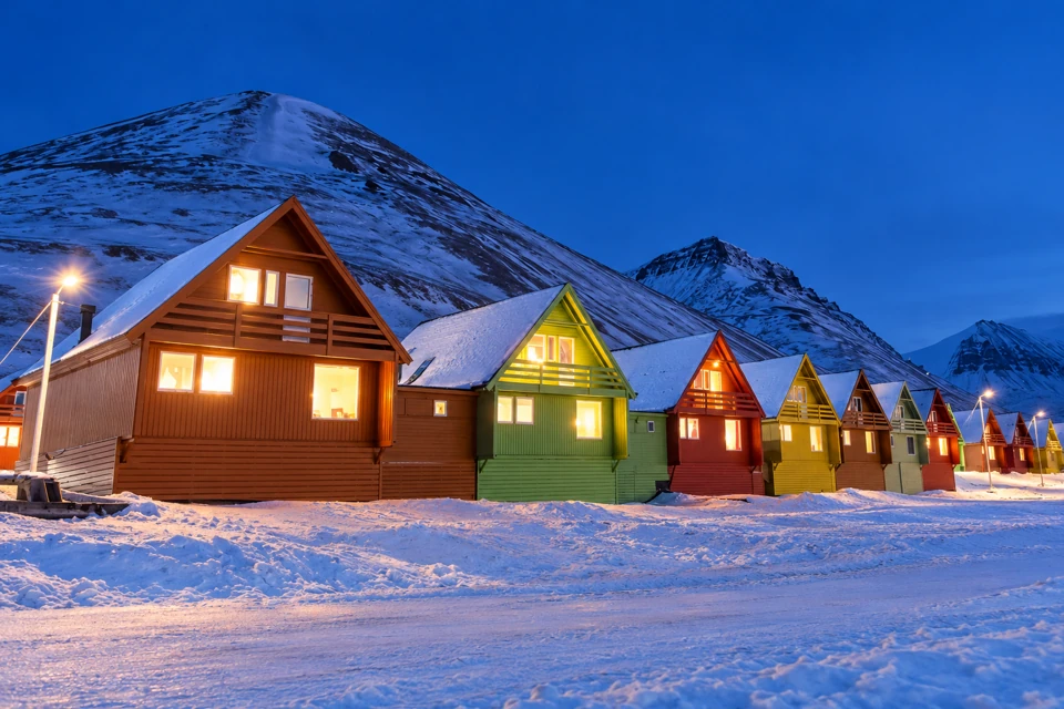 Colorful pitched-roof houses under a snowy mountain ridge in Longyearbyen at blue twilight.