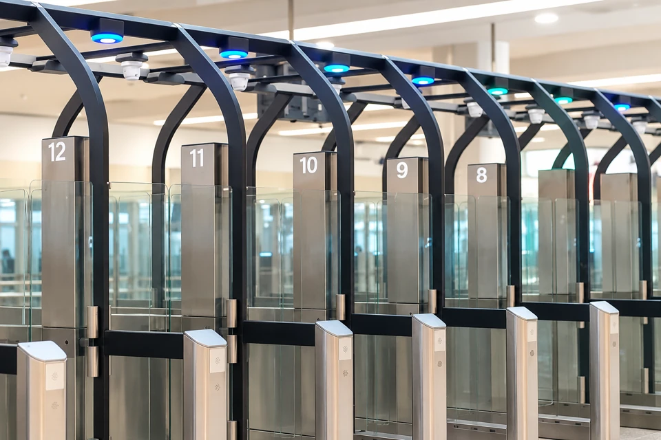 INM immigration gates at Cancun Airport, with dark angular frames, clear glass panels, silver pedestals and cyan status lights.