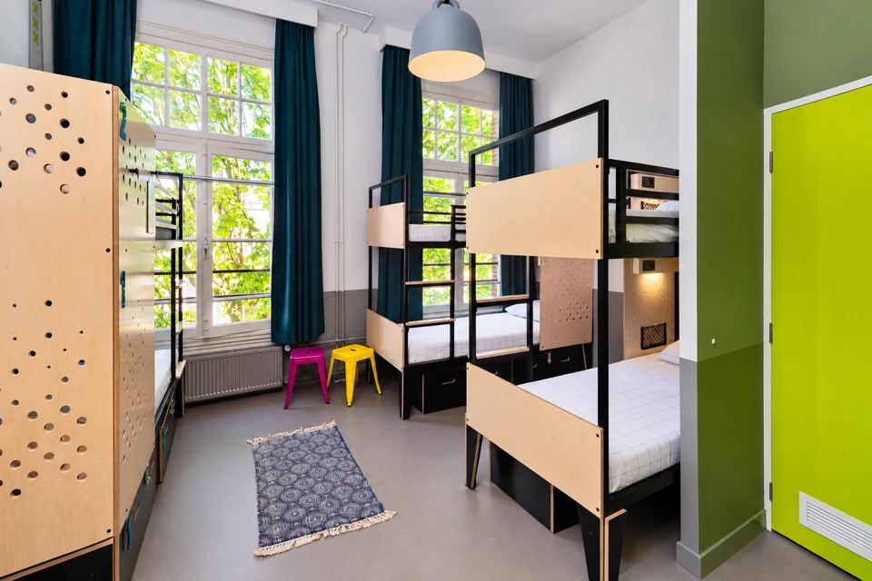 A compact shared hostel dormitory in Amsterdam, with two-level black steel and pale plywood bunks, teal curtains, tall windows and a lime-green door.