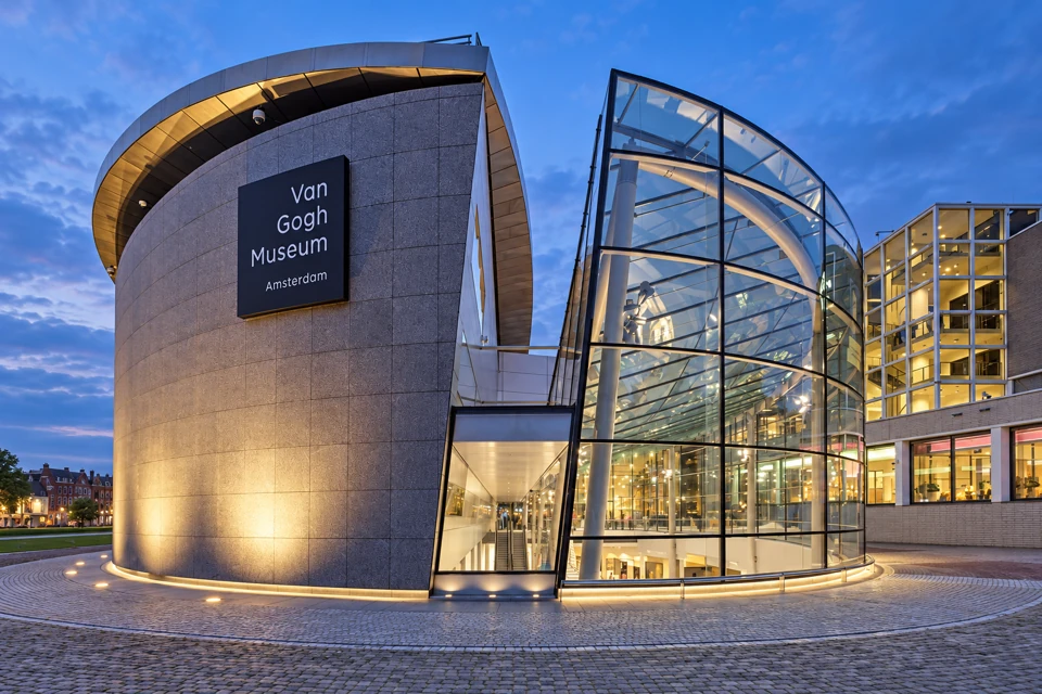 The Van Gogh Museum in Amsterdam at dusk, with a curved gray-stone facade and permanent museum sign beside its transparent glass entrance pavilion.