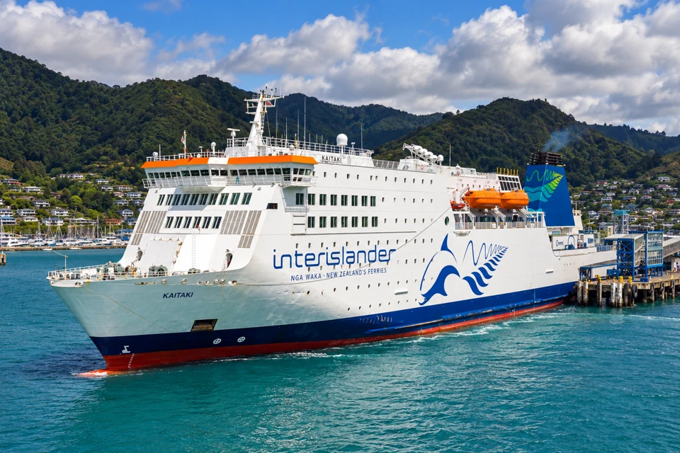 Interislander's Kaitaki ferry with a white hull, blue fern emblem, orange lifeboats and blue-green funnel beside a New Zealand terminal.