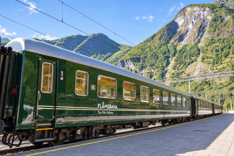 A dark green Flåmsbana carriage beside an empty platform below the steep green and rocky Flåm valley.