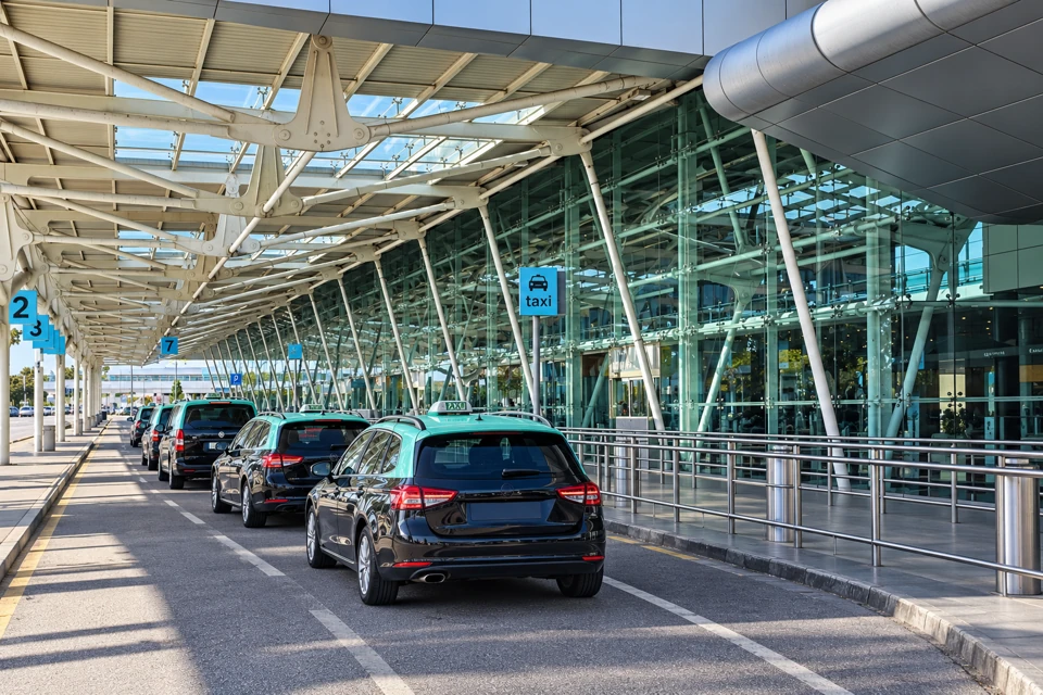 Black taxis with turquoise-green roofs beneath the white steel canopy at Lisbon Airport Terminal 1 arrivals.