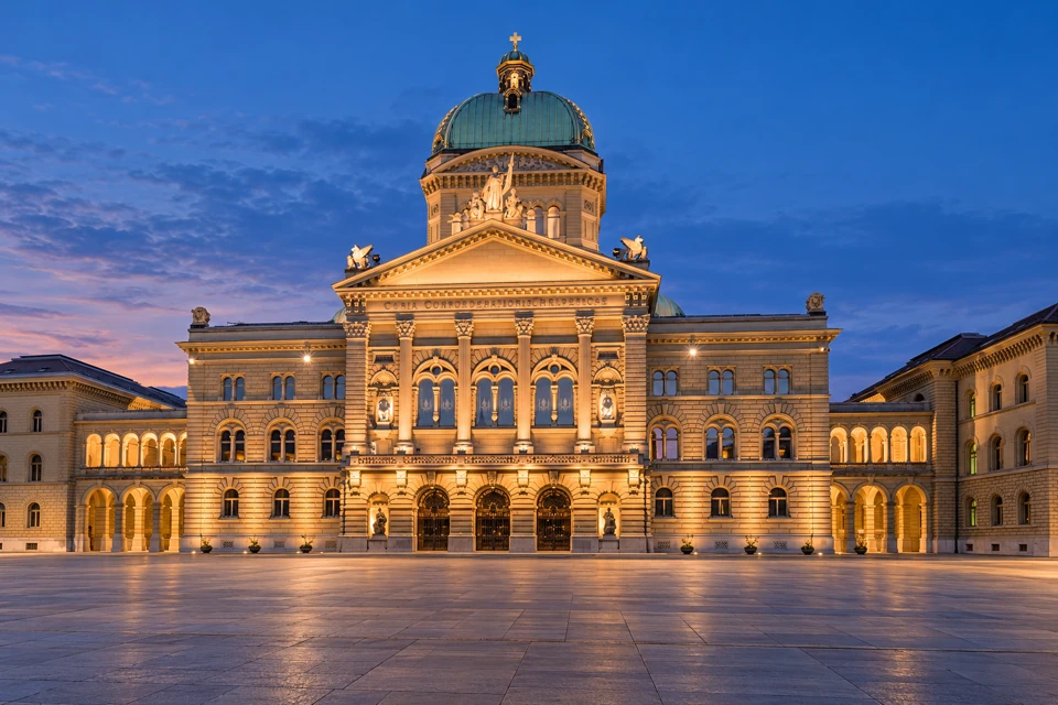 The Federal Palace in Bern, with its copper-green dome, columned sandstone facade and illuminated arcades facing Bundesplatz at blue hour.