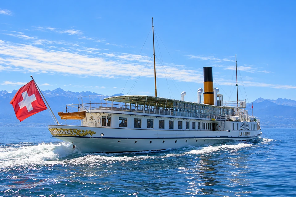 CGN's La Suisse paddle steamer on Lake Geneva, with white decks, a yellow-and-black funnel, gold stern scrollwork and a Swiss flag.