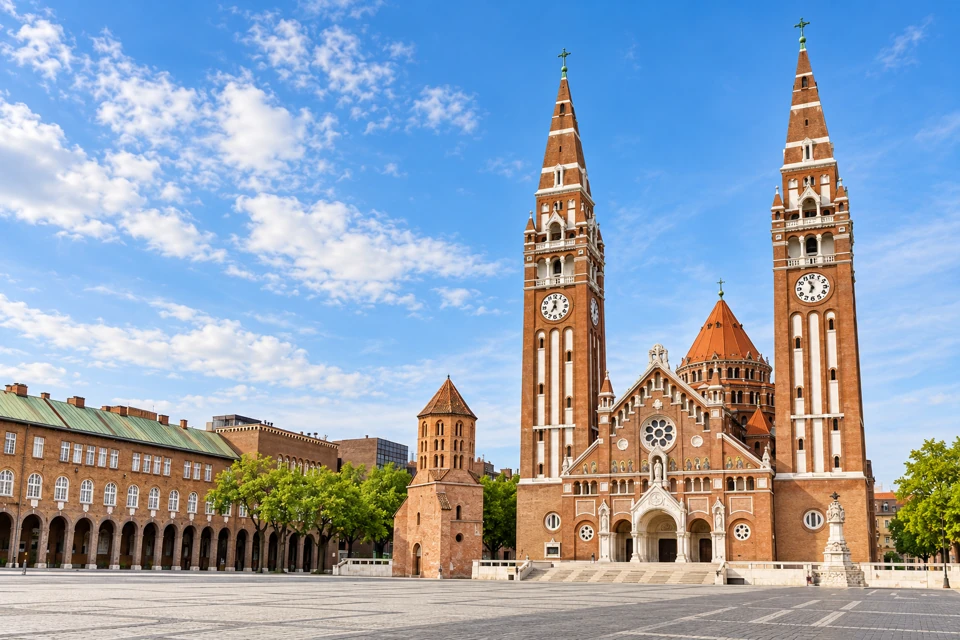 Szeged's Votive Church with twin brick spires, the smaller Domotor Tower and arcaded buildings around Dom Square.