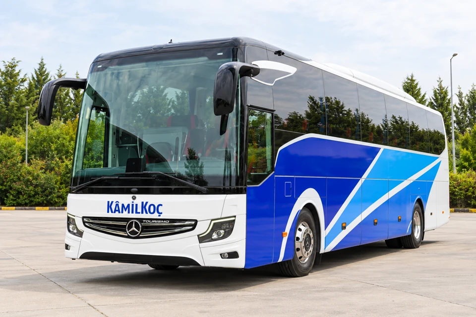 A Kamil Koc Mercedes-Benz Tourismo coach with white front, cobalt-blue side panels and diagonal white bands.