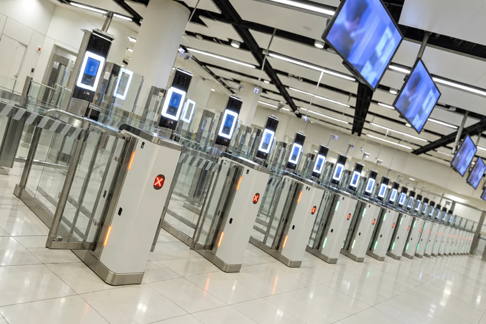 A bank of UK ePassport gates with silver pedestals, clear glass lanes, passport readers and overhead blue screens.