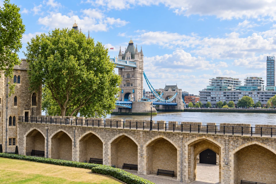 The Tower of London's stone wall walk above pointed arches and empty benches, with the River Thames and Tower Bridge behind.