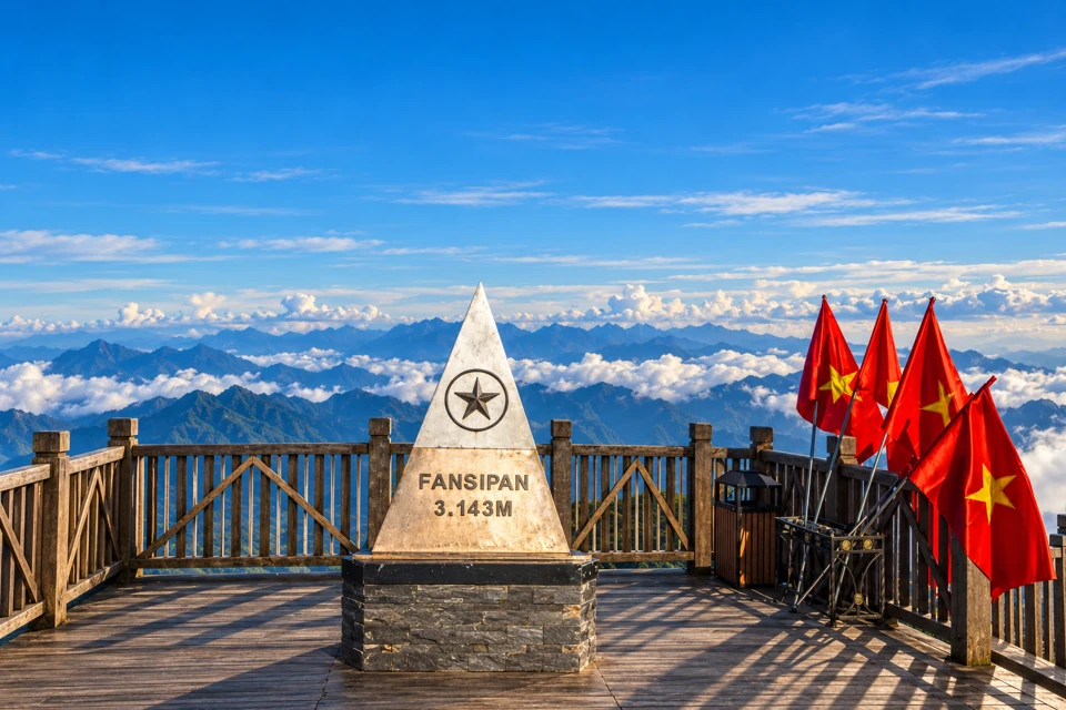 The triangular Fansipan summit marker on a wooden viewing deck above mountain ridges and clouds.