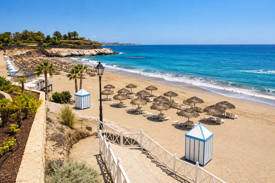 Pale sand and straw parasols at Playa del Duque sit below a palm-lined promenade, with blue-and-white changing booths and turquoise water beside the rocky Adeje headland.