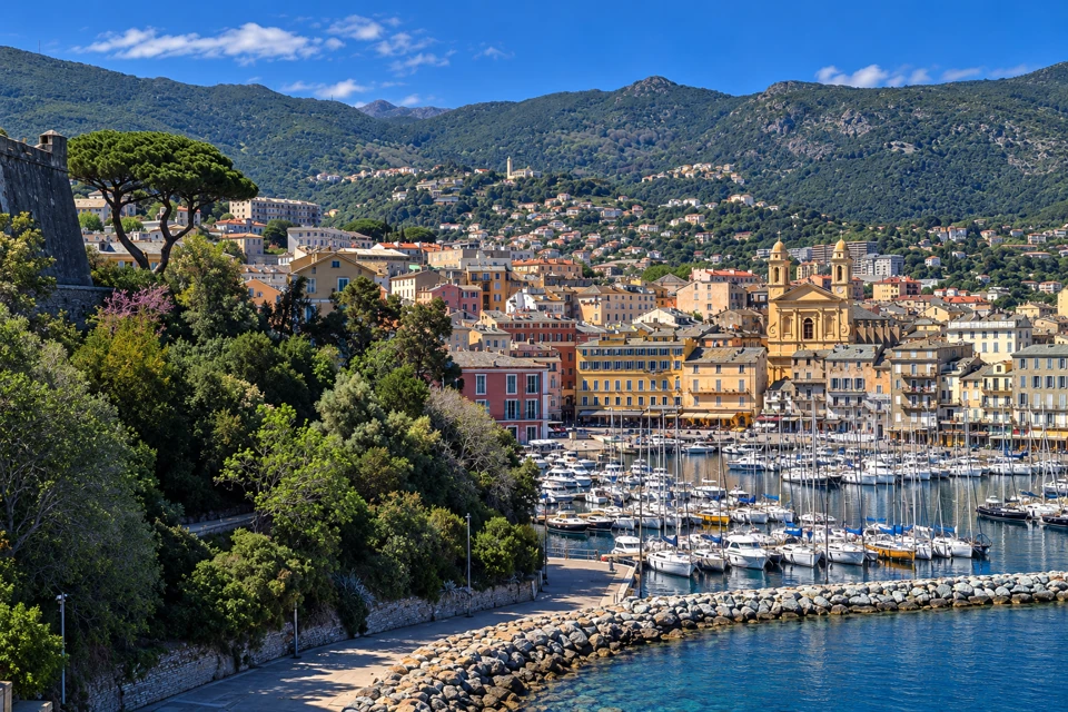Bastia's marina and pastel waterfront buildings sit beneath the twin bell towers of Saint-Jean-Baptiste, with wooded Corsican hills behind.