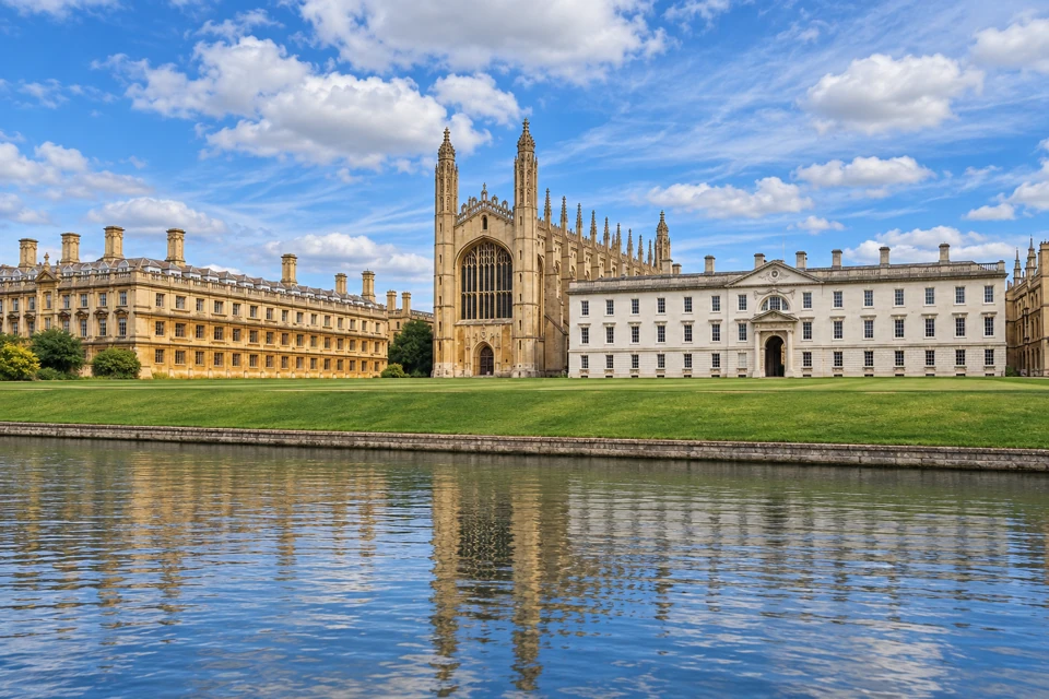 King's College Chapel's Gothic west window and slender pinnacles beside college buildings, lawn and a quiet River Cam in Cambridge.