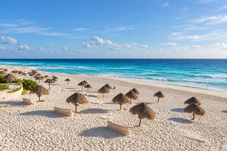 White sand, scattered thatched palapas and low coastal plants at Cancun's Playa Delfines beside the turquoise Caribbean Sea.