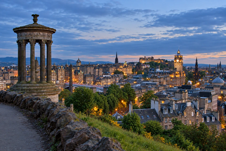 The circular Dugald Stewart Monument on Calton Hill overlooks Edinburgh's stone skyline, the Balmoral clock tower and Edinburgh Castle at dusk.