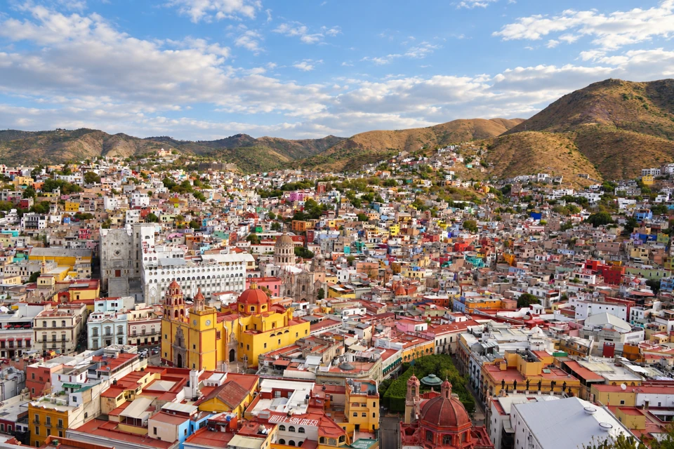 Guanajuato's yellow basilica with red dome stands among tightly packed colorful buildings below brown-green hills.