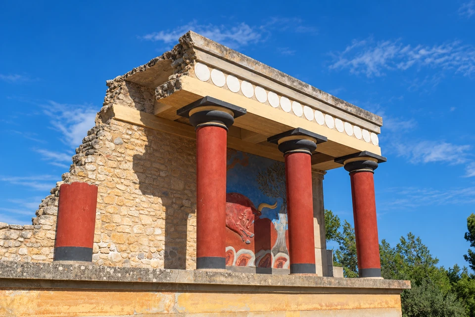 Three full red columns with dark capitals support the reconstructed north entrance at Knossos, with a bull fresco behind them and a broken column at the left.