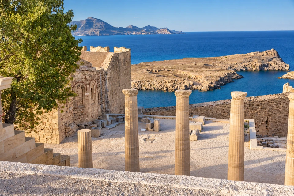 Freestanding pale columns at the Lindos Acropolis overlook a blue coastal bay, with an old stone wall and leafy tree at the left.