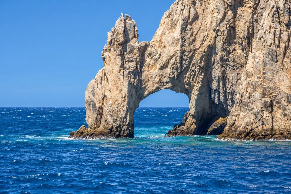 The pale weathered rock arch of Cabo San Lucas rises from blue water, with a narrow jagged rock pinnacle beside the opening.