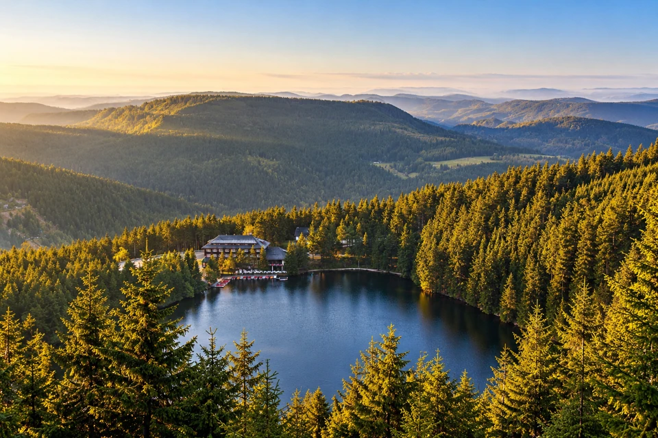 Mummelsee and its lakeside lodge surrounded by forest below layered low Black Forest ridges in soft golden light.