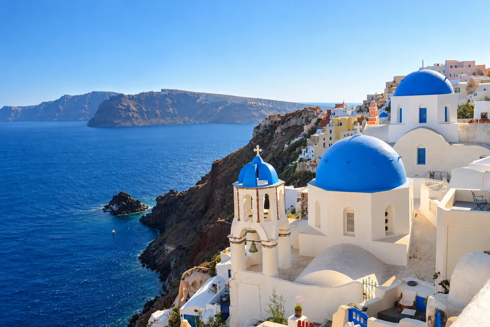 Blue-domed white churches in Oia stand above Santorini's dark caldera cliffs and deep blue sea, with the opposite island rim across the water.