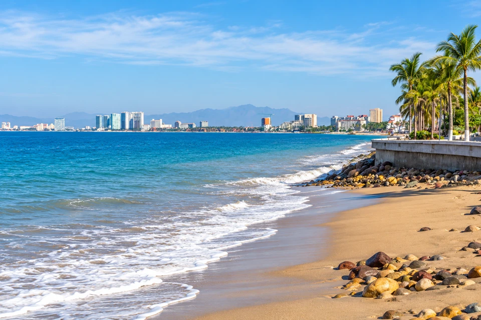Puerto Vallarta's sandy Malecon beach beside a palm-lined seawall, with the city and mountains curving around Banderas Bay.