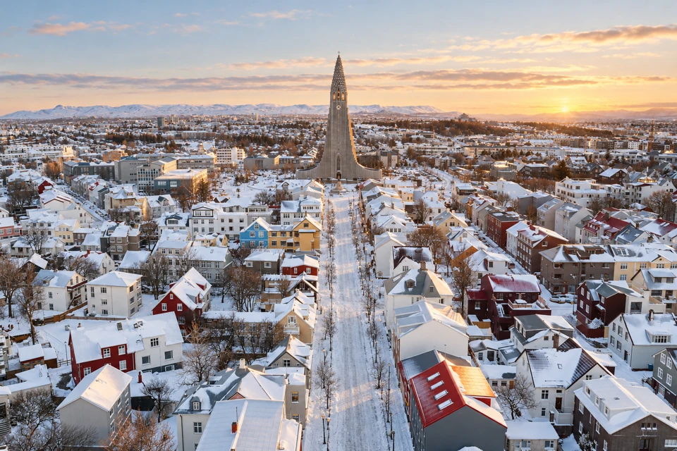 Hallgrimskirkja above the colorful low-rise roofs of central Reykjavik after snowfall, with low winter light on the horizon.